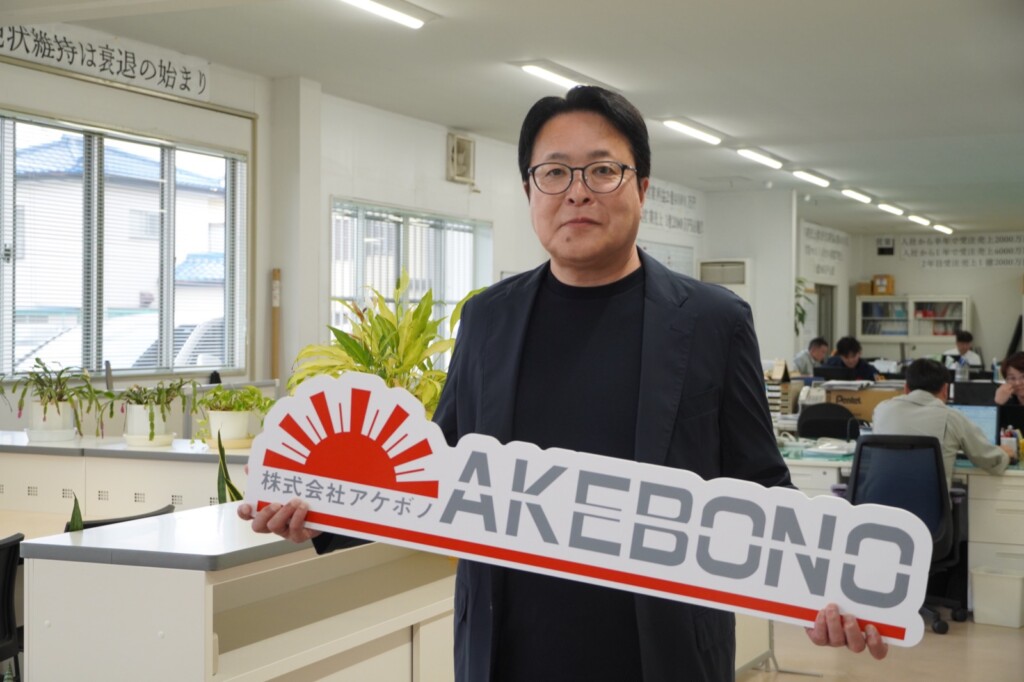 株式会社アケボノのロゴを持つ細田健一社長と本社オフィス