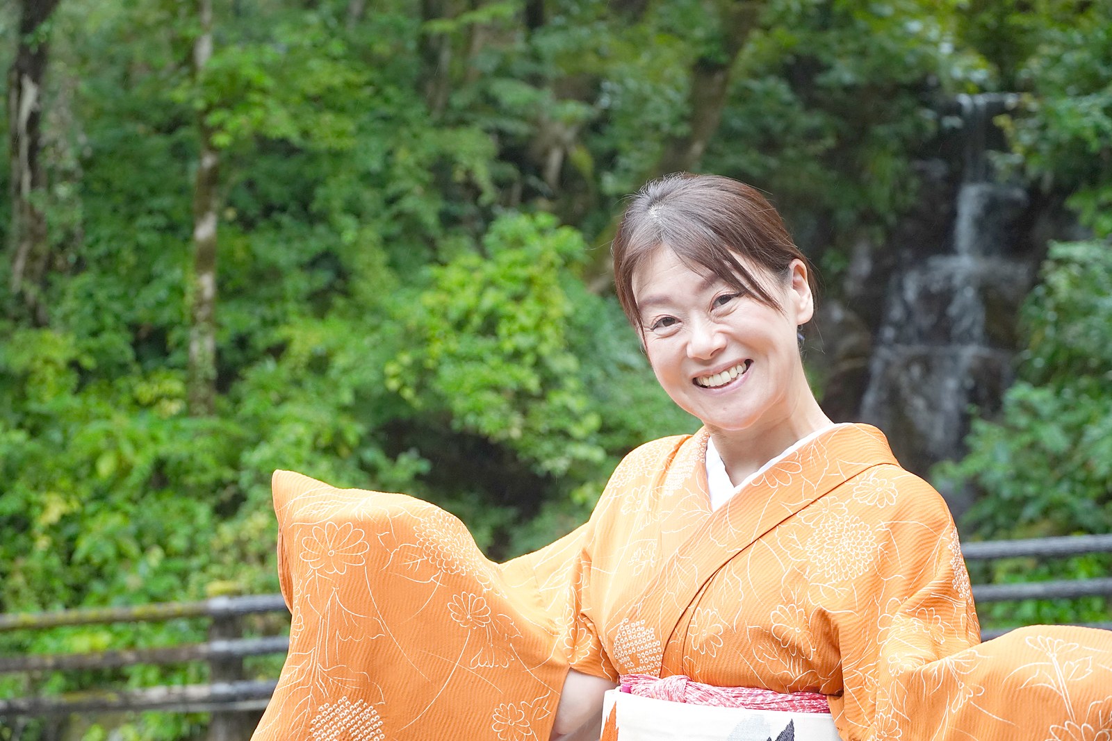 名栗の新緑を背に、着物の袖を広げて笑う名栗温泉・大松閣の女将 柏木由香さん